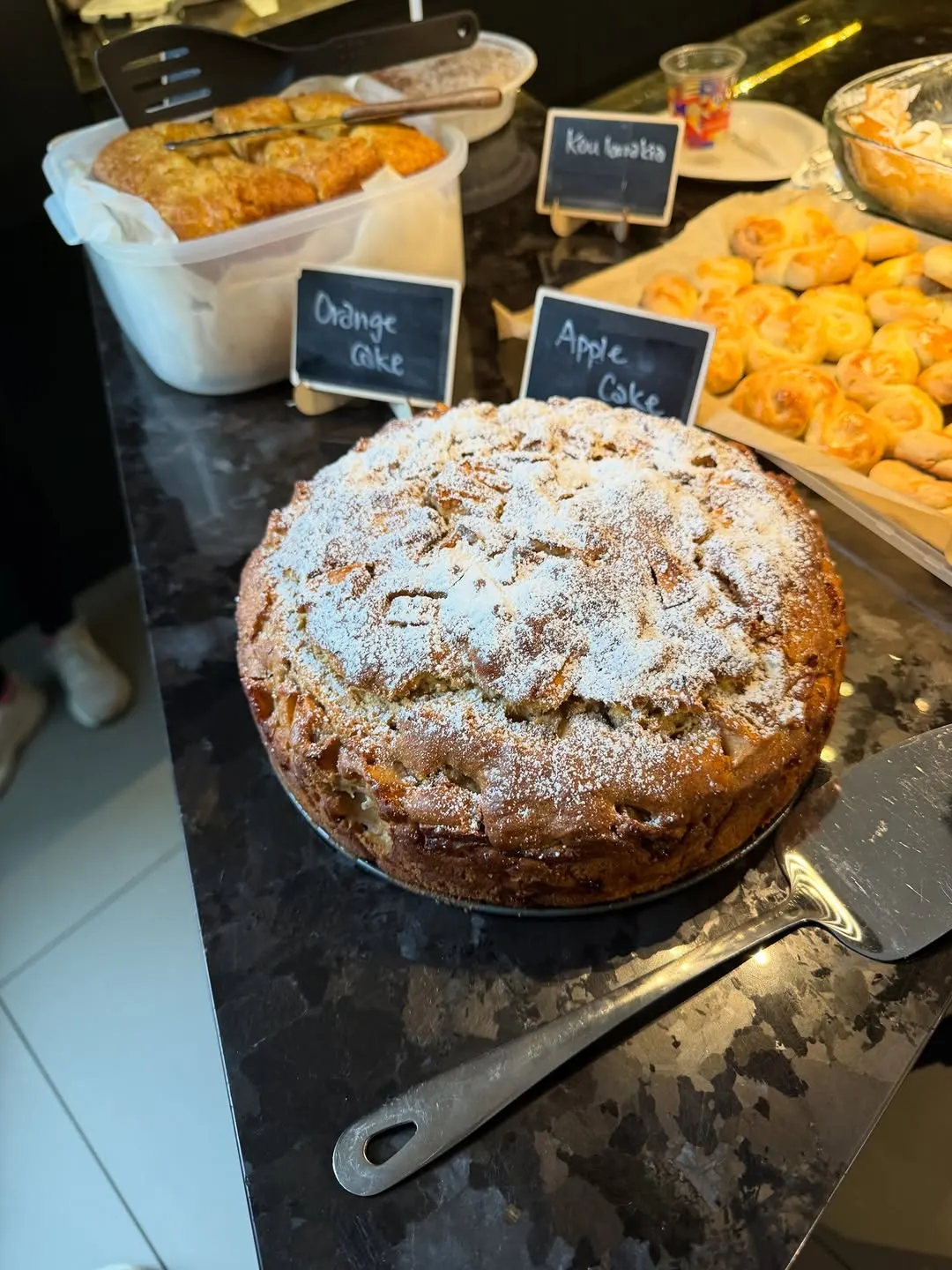 Apple cake dusted with icing sugar beside orange cake and koulourakia at the Greek night.